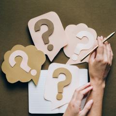 Question mark cut-outs with a hand holding a pencil and a blank notepad sitting at the centre of the image.