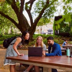 Three students sitting at wooden table outdoor looking at content on a laptop screen.
