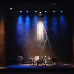Smokey and dimly lit theatre stage with several chairs and small tables.