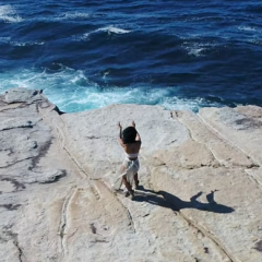 Woman standing on cliff's edge with her arms extended towards the ocean in front of her.