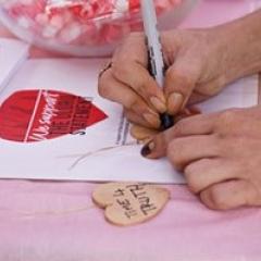 Hands holding a permanent marker as they write something on a small heart-shaped wooden cut out.