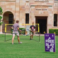 Two male students playing with oversized tennis rackets on the Great Court grass