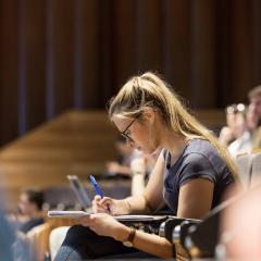 Blond woman taking notes in lecture theatre.