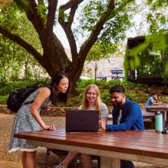 Three students sitting at wooden table outdoor looking at content on a laptop screen.
